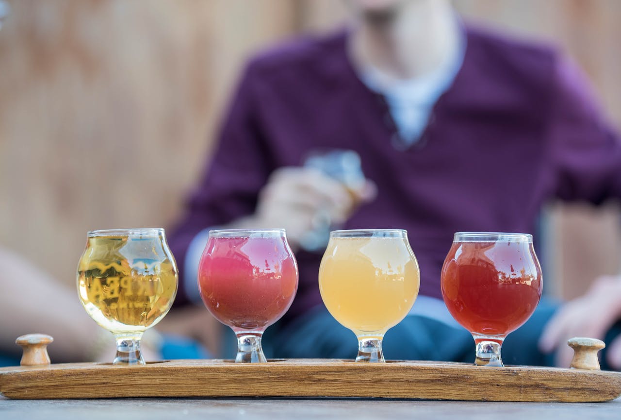 Selection of four craft beers in unique glasses on a wooden tray, with person in the background enjoying a drink.
